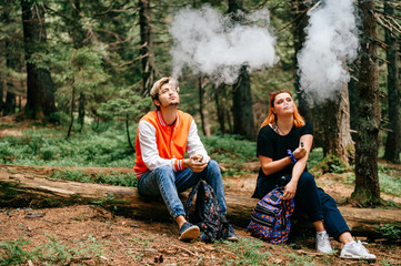 Couple of smokers enjoying electronic cigarette taste in mountain forest. Tired travelers take a rest. Sitting friends smoking at nature outdoor. Man and woman in camping trip. Bad habits people.