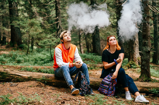 Couple Of Smokers Enjoying Electronic Cigarette Taste In Mountain Forest. Tired Travelers Take A Rest. Sitting Friends Smoking At Nature Outdoor. Man And Woman In Camping Trip. Bad Habits People.