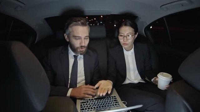 Medium shot of bearded businessman in suit and Asian businesswoman in glasses sitting in backseat of moving car and discussing work while looking at laptop