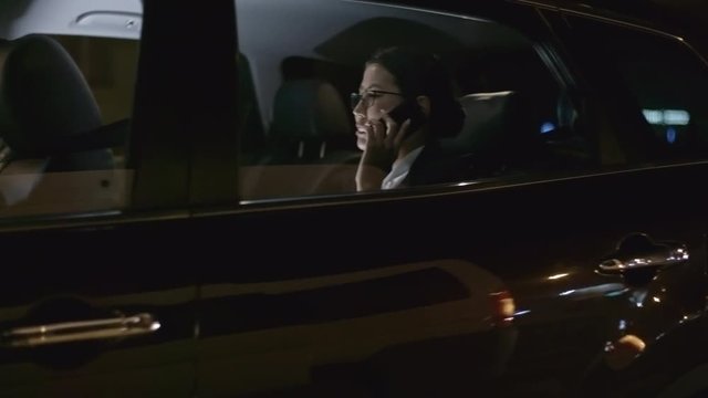 Tracking with side view of Asian businesswoman in glasses talking with business partner on mobile phone while riding in backseat of car at night