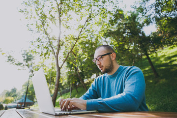Fototapeta premium Young successful smart man businessman or student in casual blue shirt glasses sitting at table with mobile phone in city park using laptop working outdoors on green nature. Mobile Office concept.