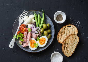 Healthy breakfast or snack - plate of canned tuna, green beans, mozzarella cheese, tomatoes, boiled egg, olives, grilled bread a dark background, top view