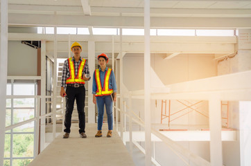 asian couple of architect, engineer or technician man and woman with safety helmet holding blueprint standing under roof in construction site building to greeting start up project, industry concept