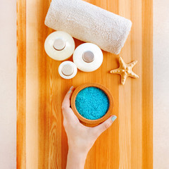 Plakat Top view of hand, taking a bowl with blue sea salt scrub, standing on wooden table with towel and bottles of spa cosmetic.