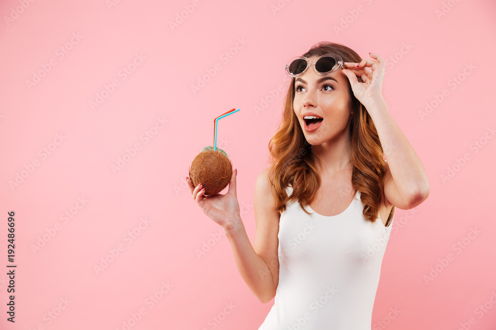 Sticker fascinating brunette woman in swimsuit and sunglasses smiling and looking aside while holding coconu