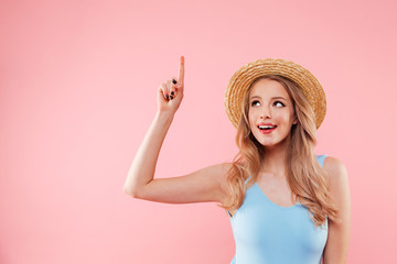 Charming young girl 20s in colorful one-piece swimsuit and straw hat smiling and pointing finger...