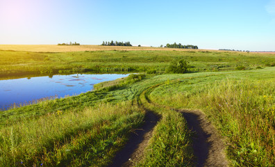 Fototapeta na wymiar Sunny summer landscape with ground country road.Field of golden ripe wheat.Green hills with growing trees.River Upa in Tula region, Russia.Countryside scene.Panoramic view.