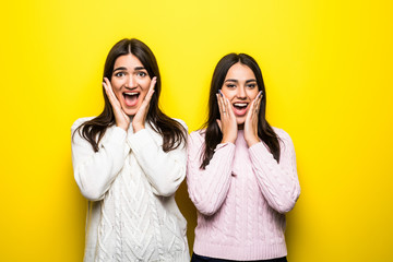 Portrait of two excited girls dressed in sweaters screaming and looking at camera isolated over yellow background