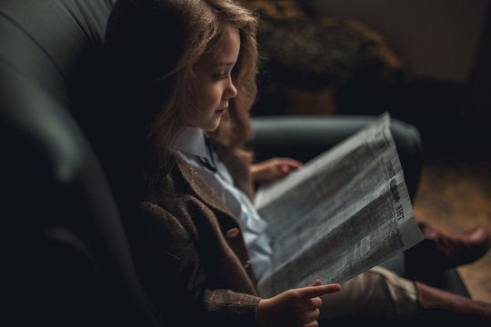 Child girl sits in armchair and reads newspaper.