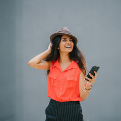 Image of an attractive and young Indian lady is taking a selfie while laughing against a plain background. She is using her smartphone and wearing fedora hat with her  very casual outfit.