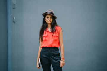 Fototapeta premium Portrait of a fashionable, young and attractive Indian woman standing outdoors against a blue grey background and looking up. She is dressed fashionably with a hat, holding a notebook. She looks cheer