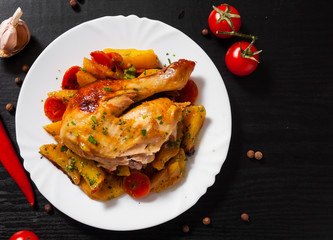 Fried chicken legs with carrots, onions and potatoes served on a white plate on dark wooden background