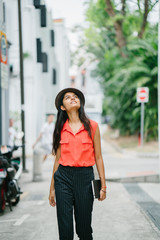 Portrait of a young Asian Indian model woman walking in the day and smiling along a street. She is dressed in smart casual clothes and a fedora hat in the summer sun in a fashionable and stylish way.