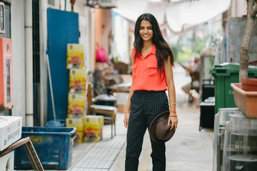 An image of an attractive and fashionably looking Indian Asian lady standing in an alley  while holding her fedora hat during the day and enjoying the backstreets of Singapore.