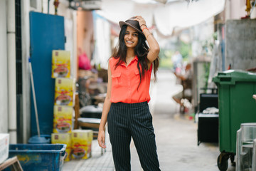 Portrait of a young Indian Asian woman standing in a back alley in Asia. She is fashionably dressed and smiling as she holds her brown fedora hat
