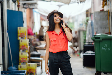 An image of a young and attractive Indian lady walking along an alley during the day. She is talking to someone on her phone and looks amazed and excited.
