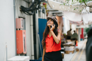 Portrait of a very fashionable and young Indian woman wandering in an alley while talking to someone on her smartphone. She is  dressed in a casual outfit and wearing a brown fedora hat for the day.