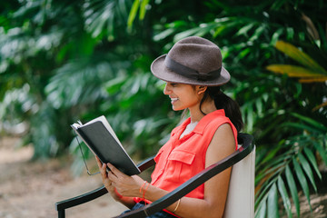 Portrait of a young and attractive Indian Asian girl reading a book in the sun during the day while sitting in a chair in a green park. She is smiling and enjoying herself and is fashionably dressed.