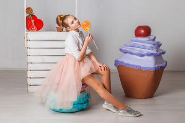 Beautiful teenage model girl in pink tutu or tulle skirts with colorful yellow lollipops in studio on white background  with artifitial big macaroons, fancy cakes and sweets