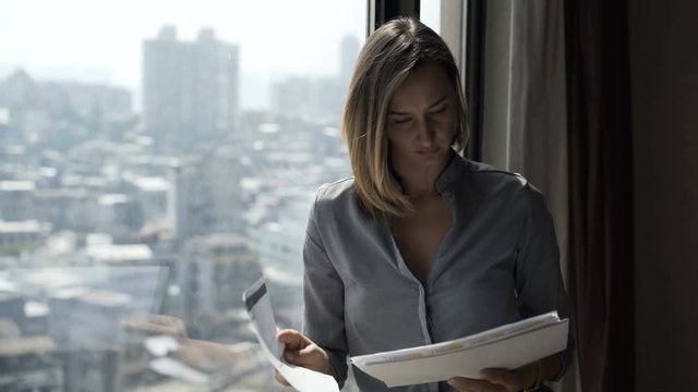 Young businesswoman reading, analyzing documents standing by window at office
