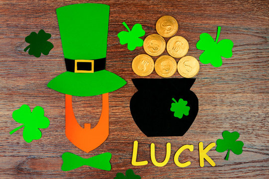 Saint Patrick's Day. Gold wooden letters "luck" with silhouette of leprechaun in green hat and green tie bow with green three petal clover, pot with gold coins on wooden background