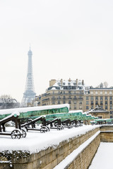 Winter in Paris in the snow. The row of bronze cannons of the Place des Invalides with a typical parisian building and the Eiffel tower disappearing slightly in the mist in the background.