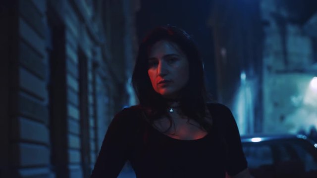 Daring attractive young woman in a trendy black outfit against the shining mysterious city, playfully looks straight to camera. Big city life, night fun, loneliness, moody atmosphere