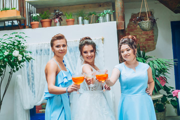 Beautiful brunette bride and gorgeous bridesmaids with bouquets posing near cozy urban space. Young woman with cocktails