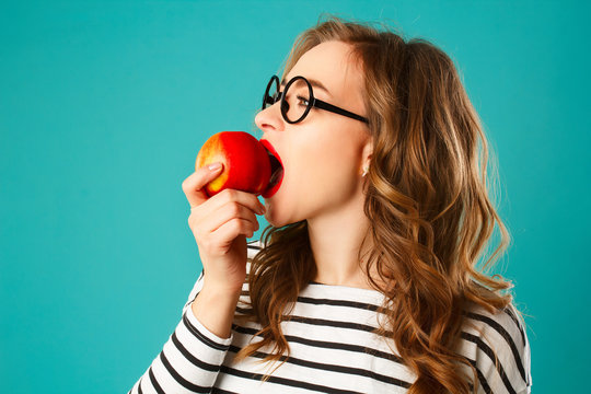 Portrait of young beautiful blond woman in round black glasses eating red apple over blue background