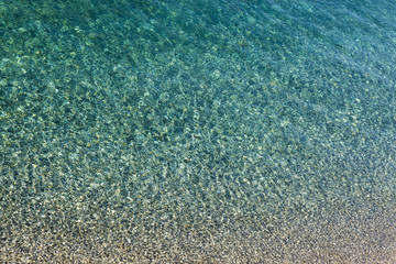 Stones and pebble under blue, azure and green sea water surface with ripple and glare of the sun as...