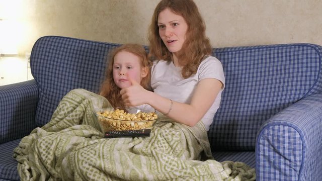 Happy loving family. Mother and her daughter child girl are eating popcorn on the bed in the room. front of the TV