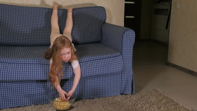 A little girl is sitting on the couch watching TV and eating popcorn
