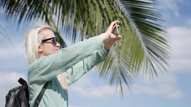 Beautiful slim woman with long blonde hair in sunglasses and green shirt standing near palm tree and take a pictures by smartphone on a blue sky background.