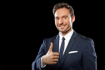 Handsome smart businessman in a classic office suit and tie showing an okay sign with thumbs up isolated on a black background, a guy you can trust