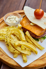 A huge juicy burger in a thin homemade pita bread with a sheet of green lettuce, melted cheddar cheese, veal cutlet and cherry tomato. Cheeseburger with French fries and white sauce on a wooden board.