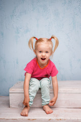 Fototapeta premium Little funny blue-eyed girl child blonde with a haircut two ponytails on her head sitting on a gossip on the background of an old textured wall in blue. Dressed in stylish bright clothes