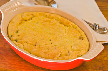 Close up, top view of a oval, red baking dish filled with homemade, freshly baked corn pudding with cilantro, chorizo sausage and an artisan fork on a yellow napkin