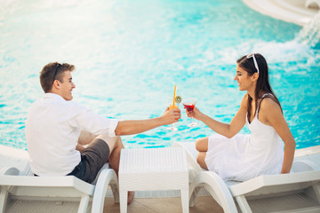 Positive happy couple having a romantic afternoon by the pool in luxury summer vacation resort.Drinking cocktails.Relaxing and enjoying spa wellness weekend.Travel, honeymoon,relationship concept
