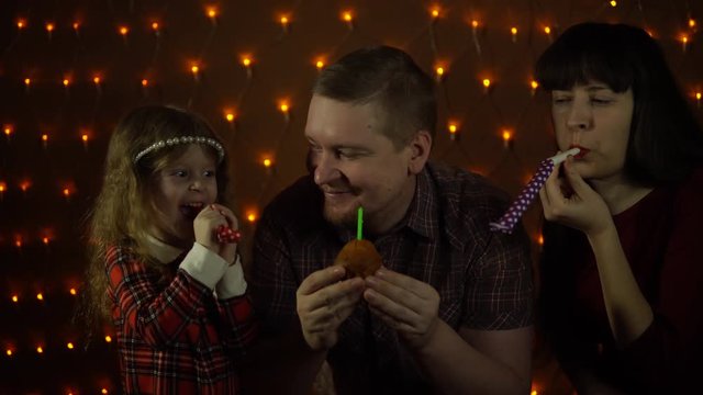 A young man blows a candle on a cupcake, his wife and daughter blowing in colorful party horns. Family together celebrate the holiday.