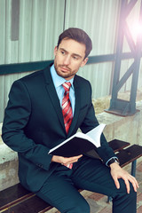 Handsome young business man with a beard in a nice suit holding a book standing on a train station...