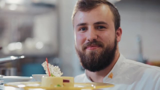 Attractive bearded confectioner finishes the preparation of dessert, puts on the top of vanilla mousse pastry white chocolate garnish decoration, then charmingly smiles straight to camera