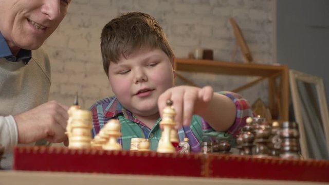 Grandfather and grandson plays chess. Elderly man teaches chess to play a young fat child. Home comfort, family idyll, cosiness concept, difference of generations, close up. 60 fps