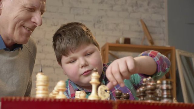 Grandfather and grandson plays chess. Elderly man teaches chess to play a young fat child. Home comfort, family idyll, cosiness concept, difference of generations, close up. 60 fps