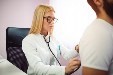 Doctor examining patient with stethoscope in medical office. Doctor using stethoscope to exam man patient heart. Portrait of female doctor examines a patient with stethoscope.