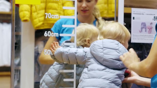 Young mother in clothing store fitting gray childrens jacket on little daughter
