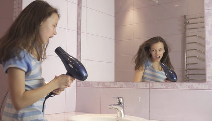 Beautiful happy girl teenager dries hair with hair dryer and sings and dances in front of a mirror in the bathroom