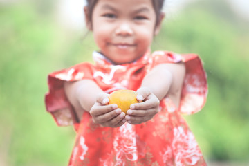 Cute asian child girl in china traditional dress holding an orange and giving you a happy in Chinese New Year