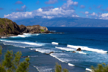 Volcanic rock, surf, and shades of the blue Pacific Ocean on the Maui coast, with the island of...