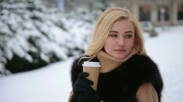 Portrait of a thoughtful beautiful girl in a beige sweater and a black coat with a Cup of coffee in a snowy city Park.