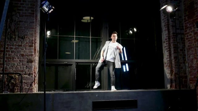 Man in black clothes dances in modern style on plinth near large window woth stage light